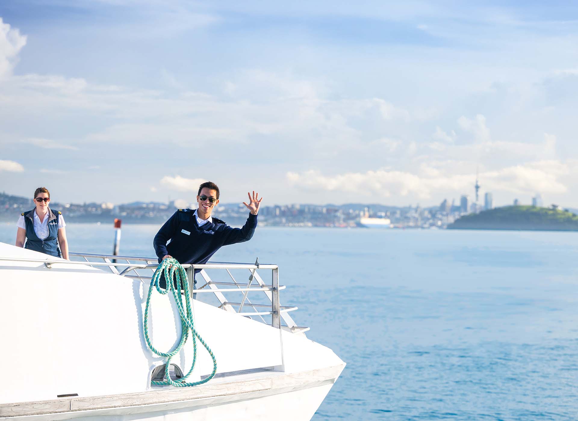 Fullers360 crew member waving from a ferry on the Waiheke Island route with the Auckland city skyline in the background - Waiheke Weekly Pass